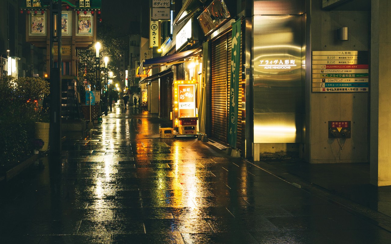 A city street at night in the rain, shop lights reflected on the wet road