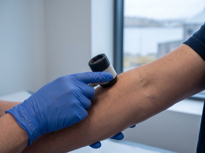A gloved hand holding a dermatoscope to a forearm